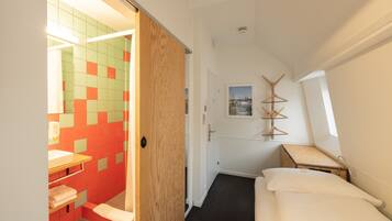 Un dormitorio con una cama, una mesita de noche de madera, una pequeña mesa, un ventanal y un baño con lavamanos y espejo.