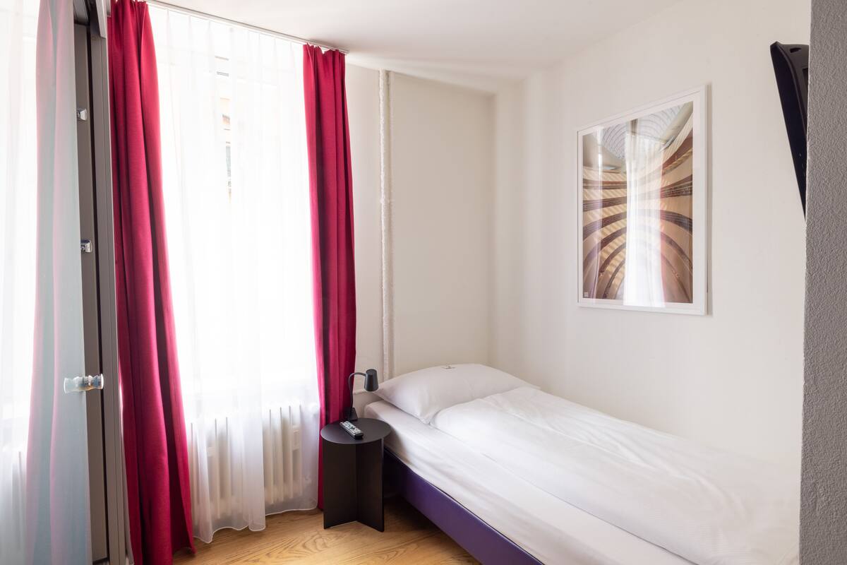Ein kleiner, sauberer Raum mit einem Bett, einem Nachttisch, einem Fenster mit roten Vorhängen und einem gerahmten Bild an der Wand.