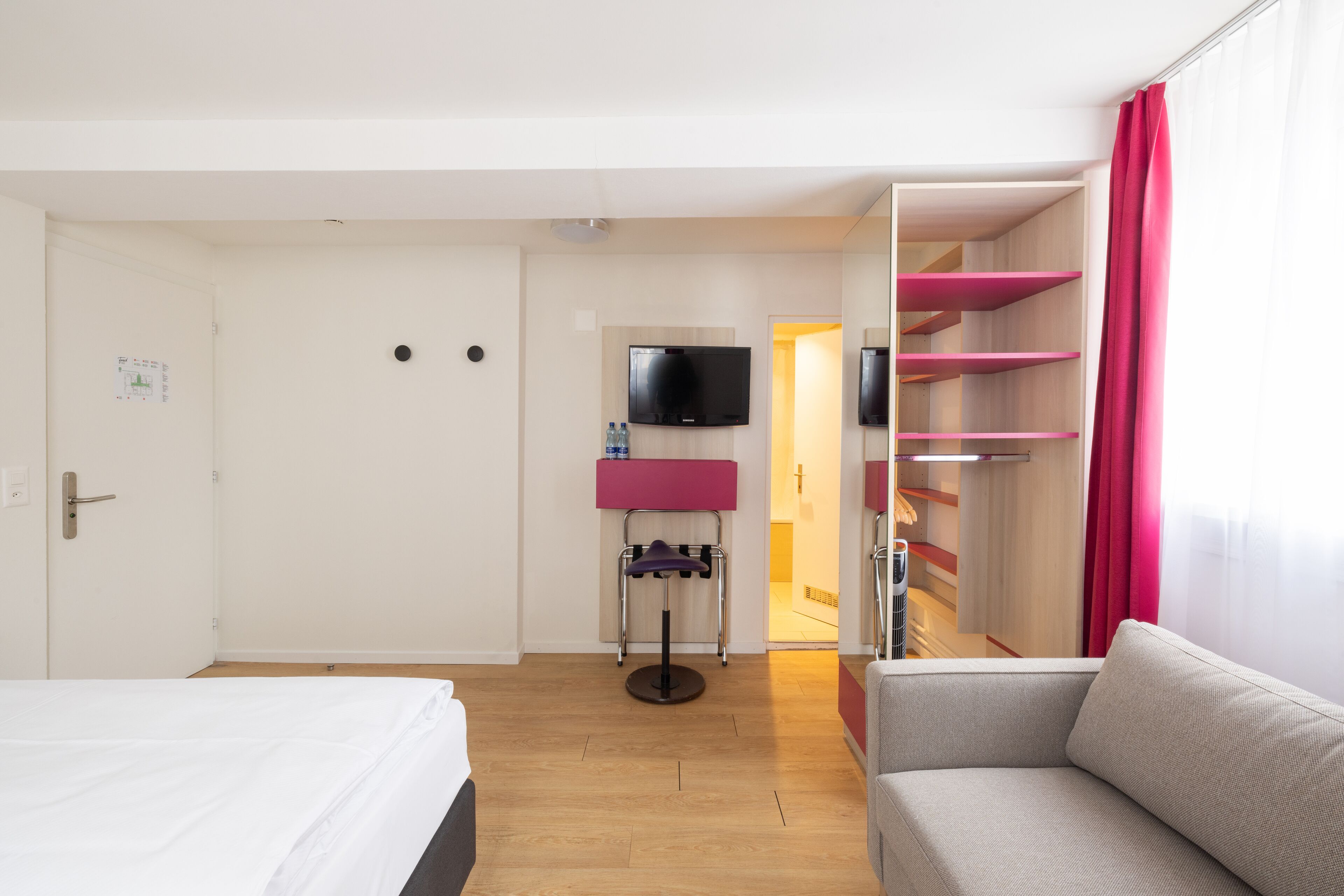 A hotel room with a bed, a sofa, a TV mounted on the wall, and a door leading to a bathroom.