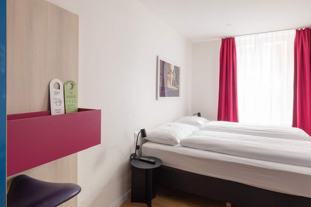 Ein modernes Hotelzimmer mit einem Bett, einem kleinen Nachttisch, einem Sessel und einem an der Wand befestigten Regal mit einem grün-weißen Schild. Ein Fenster mit roten Vorhängen rundet das Bild ab.