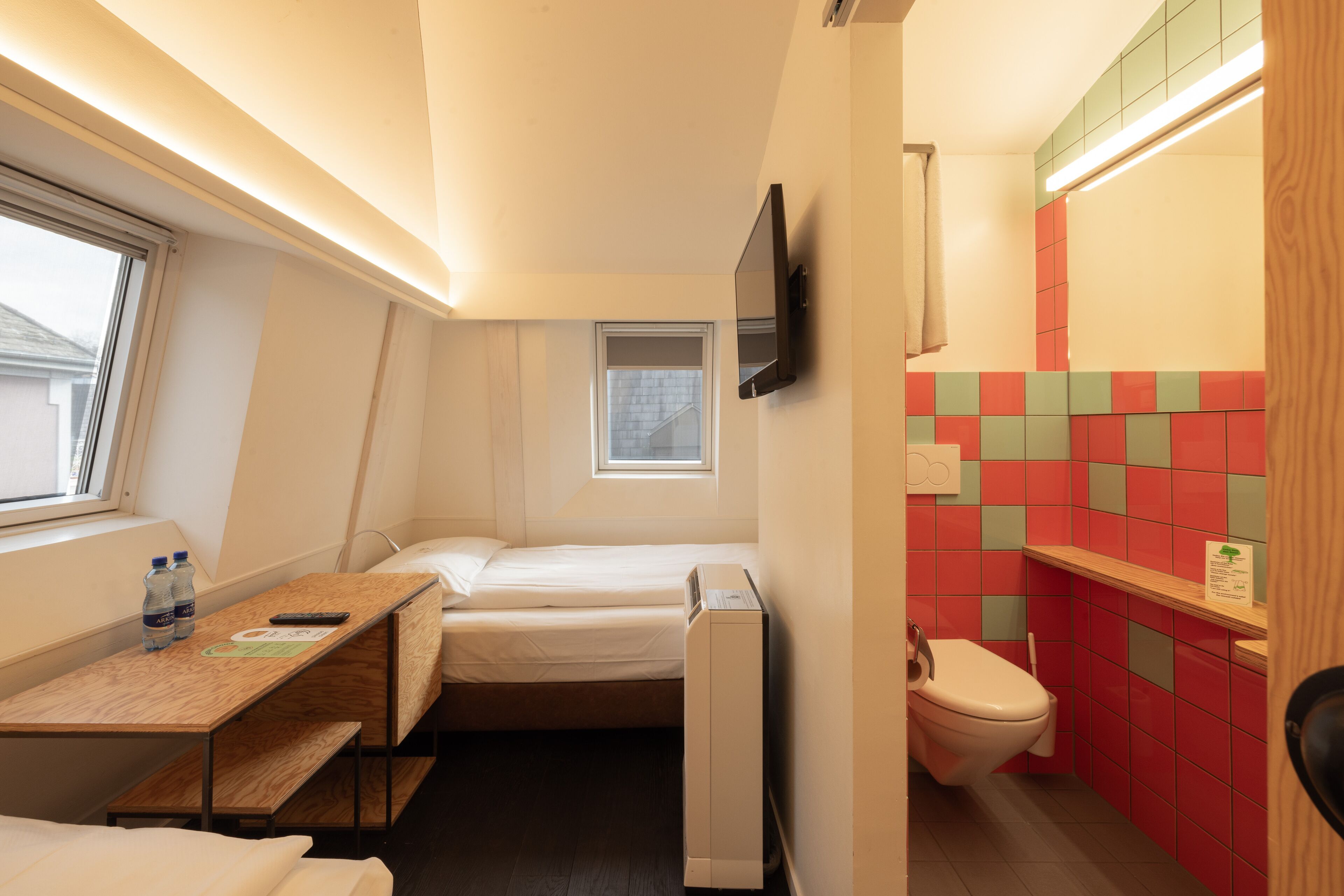 A compact hotel room with a bed, a desk with a chair, a small window, a TV mounted on the wall, and a bathroom with a toilet and a shelf.