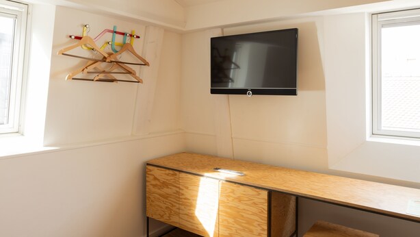 A room with a wooden desk, a TV mounted on the wall, and a window with a view of the outside. - Fred Guest House Hauptbahnhof - Self Check-in (Zürich)