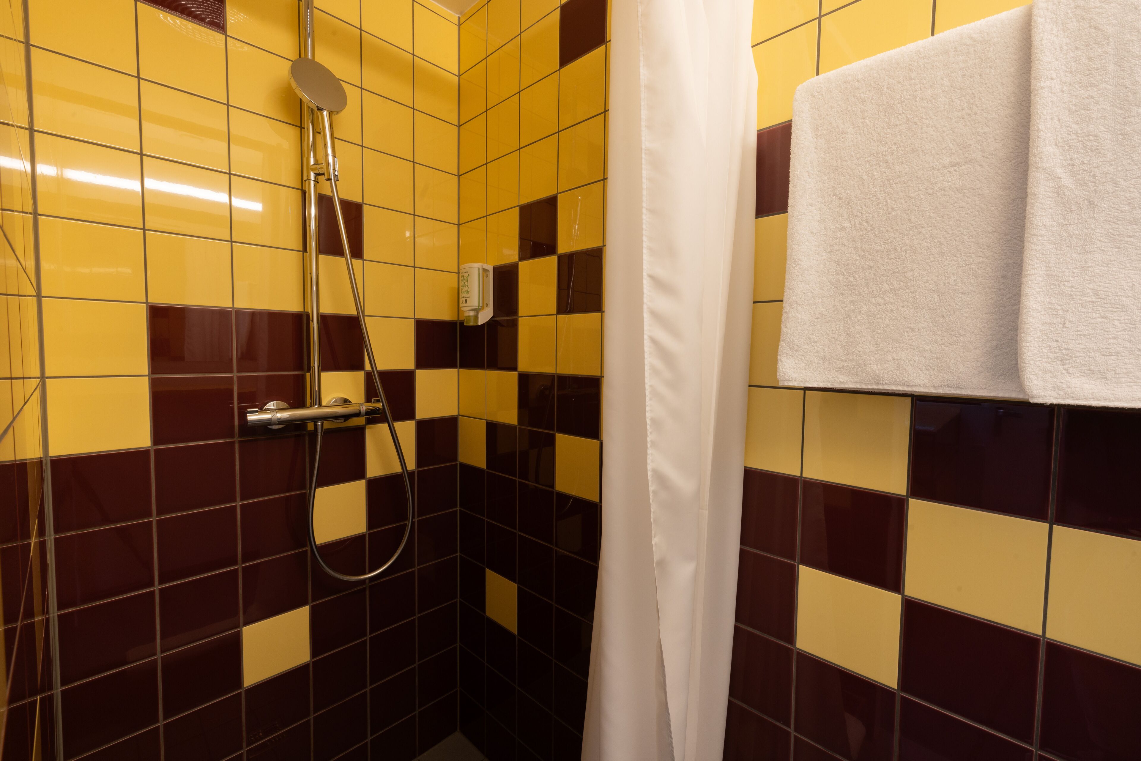Une salle de bain avec des carreaux de sol aux motifs à carreaux jaunes et bruns, une pomme de douche et un porte-serviettes avec une serviette blanche.