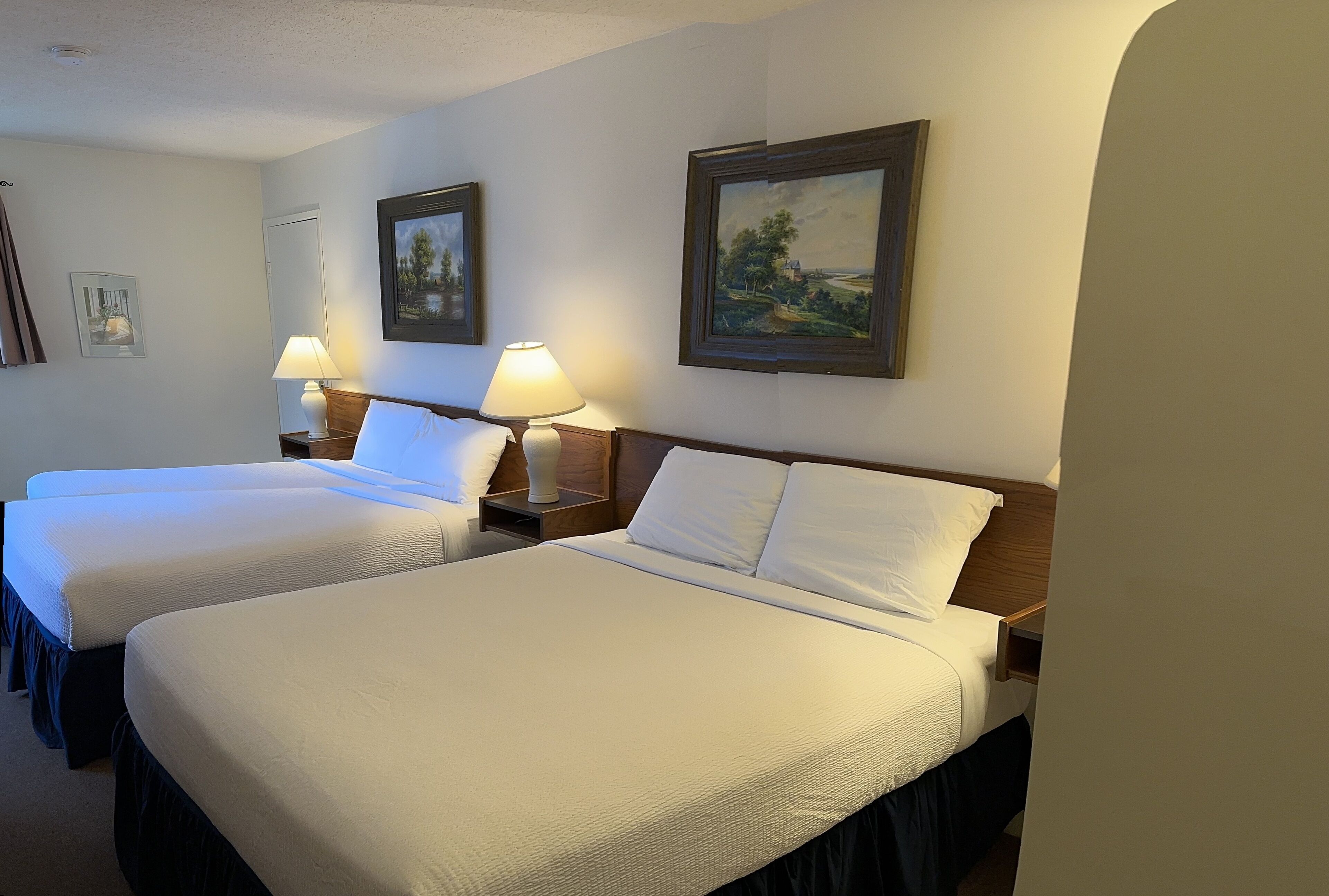 Habitación de hotel con dos camas, cada una con cabecero de madera y ropa de cama blanca. Hay dos lámparas de noche y un cuadro con un paisaje en la pared.