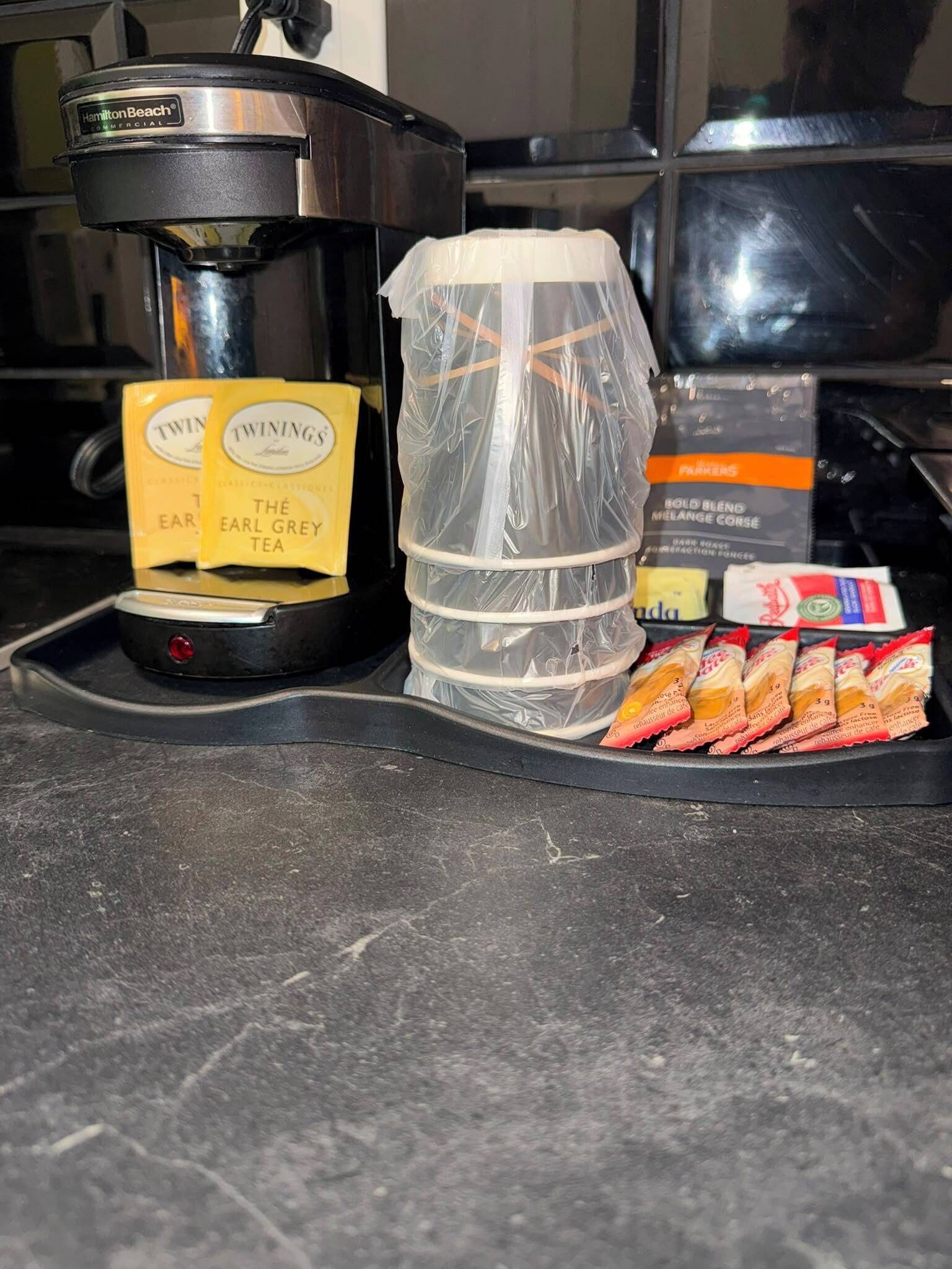 Een koffiezetapparaat, een plastic bak en een dienblad met theezakjes en snacks op een aanrecht.