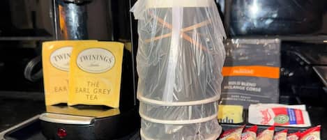 A coffee maker, a plastic container, and a tray with tea bags and snacks on a kitchen counter.