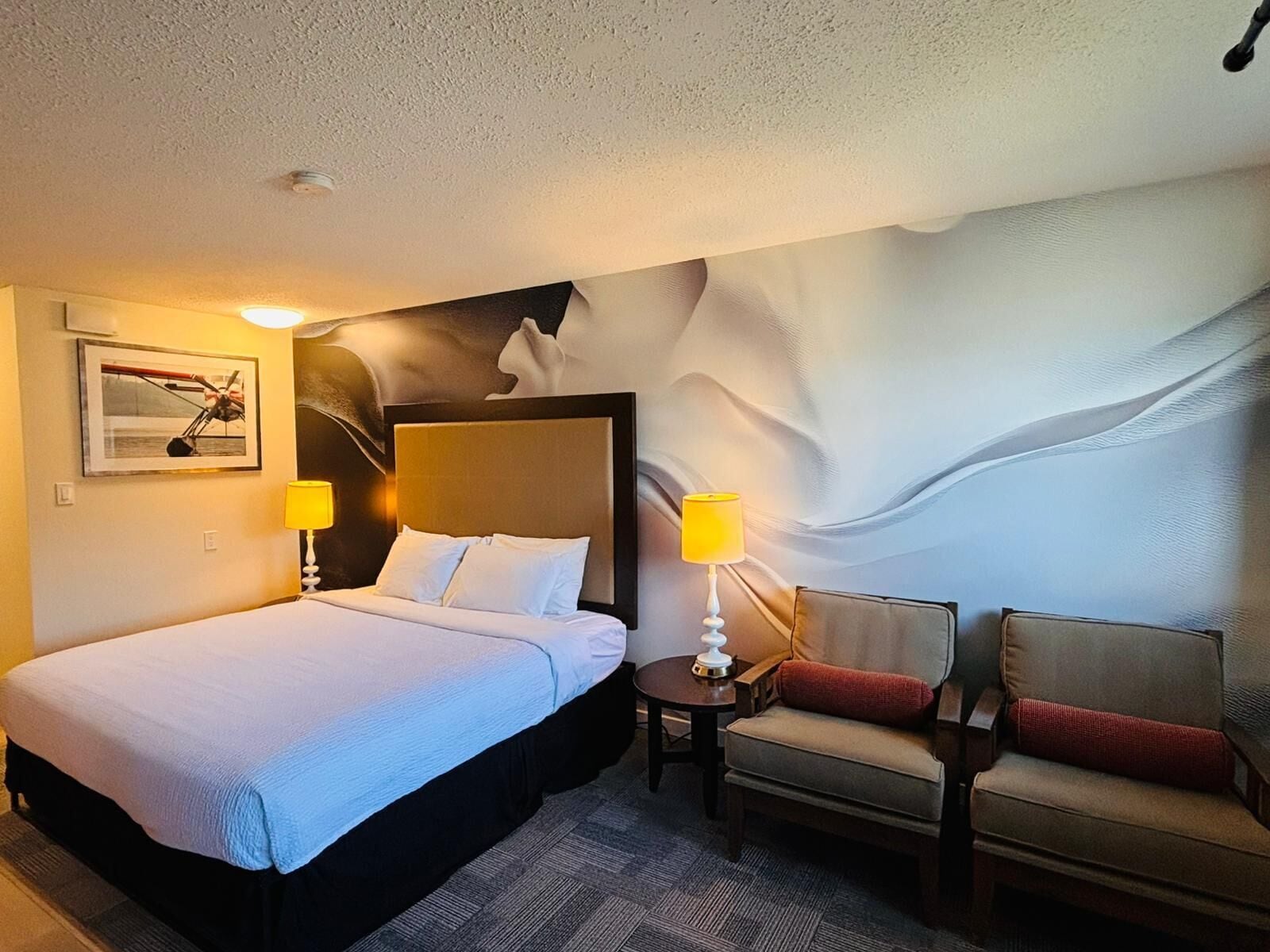 A hotel room with a large bed, two bedside lamps, a chair, a small table, and a wall mural.