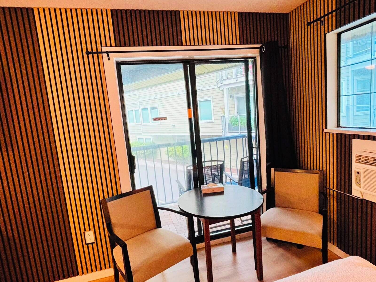 A room with a sliding glass door to a balcony, two chairs, a small table, and a window with blinds.