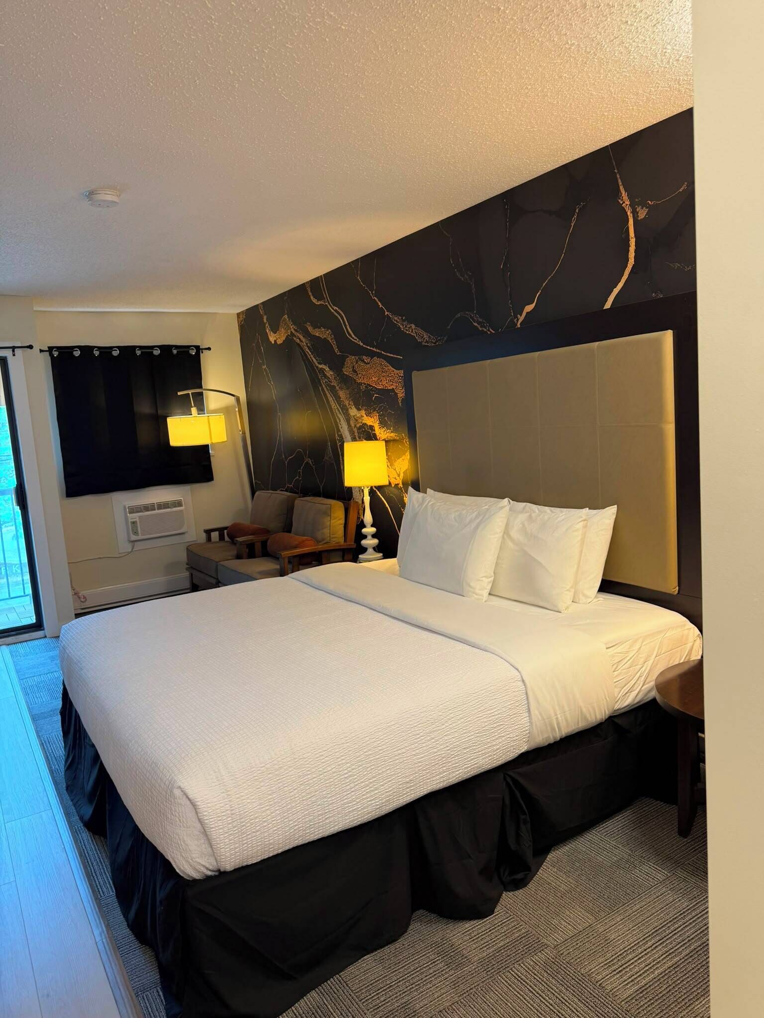 A hotel room with a large bed, a headboard, a nightstand, a lamp, a wall-mounted air conditioner, and a curtain.