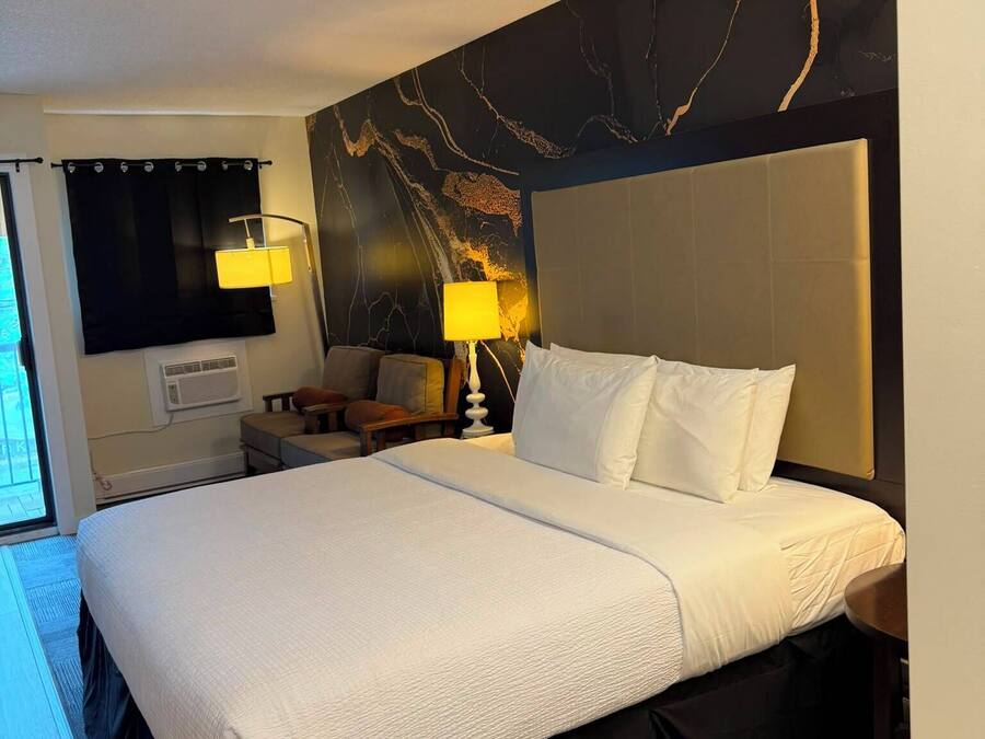 A hotel room with a large bed, a headboard, a nightstand, a lamp, a wall-mounted air conditioner, and a curtain.