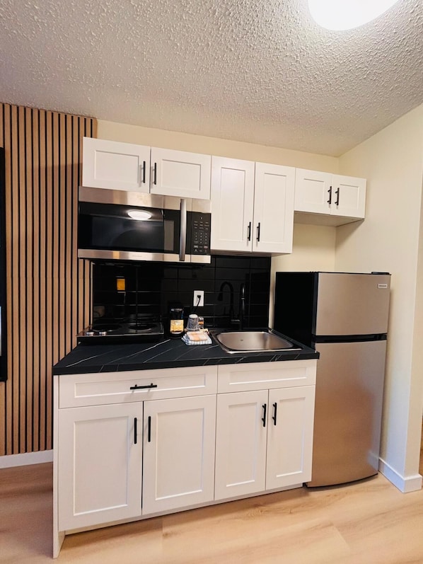 A compact kitchen with white cabinets, a black countertop, a stainless steel refrigerator, and a built-in microwave. - Wessex Inn By The Sea (Cowichan Bay)