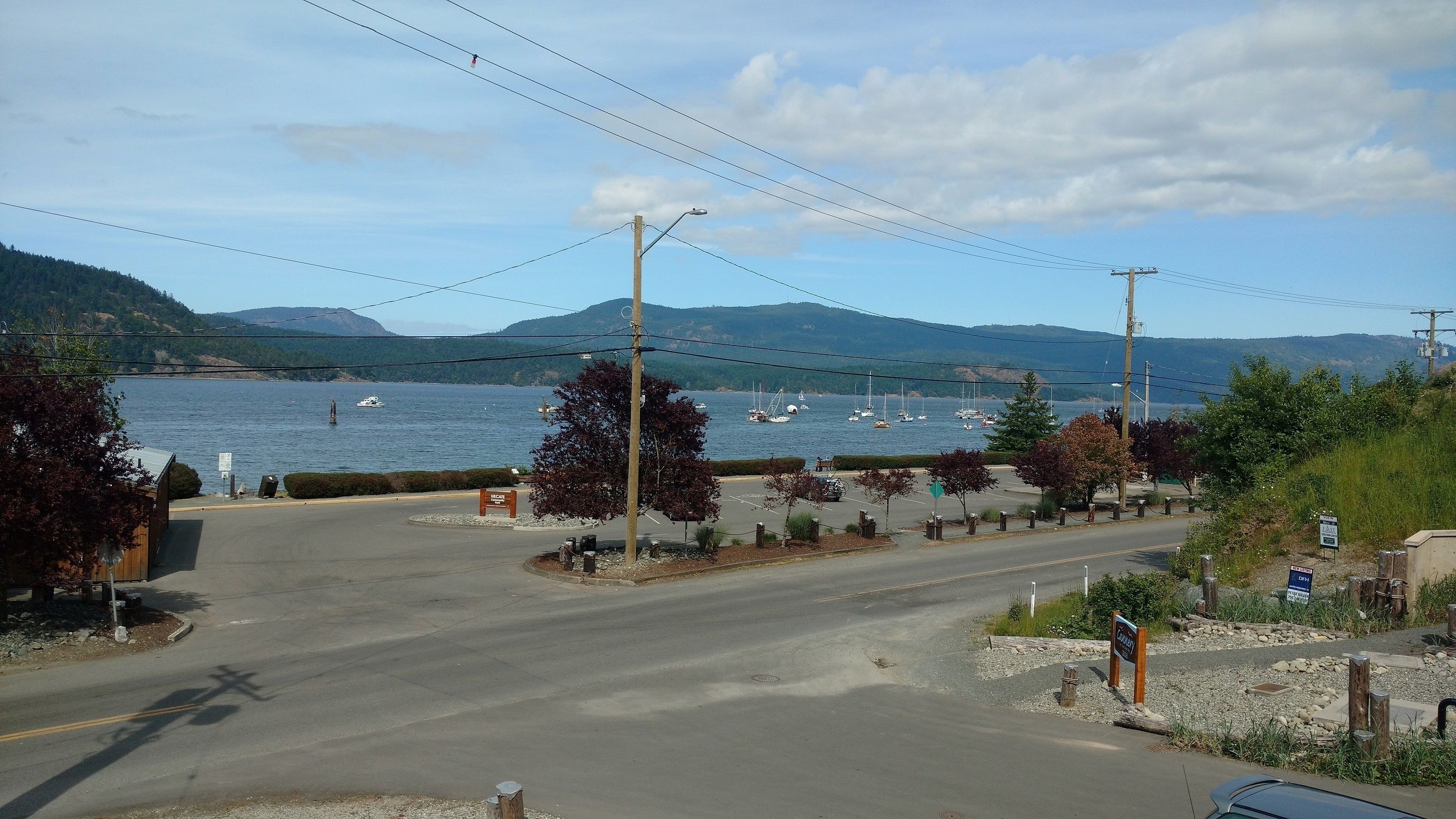 Une route côtière avec un espace de stationnement, un panneau indiquant un terrain de camping et une vue sur la mer avec des bateaux.