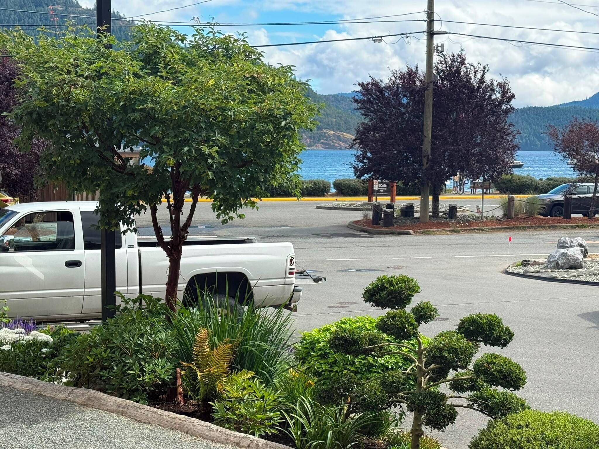 A parking lot with a white pickup truck, a tree-lined driveway, and a view of a body of water and mountains in the background.