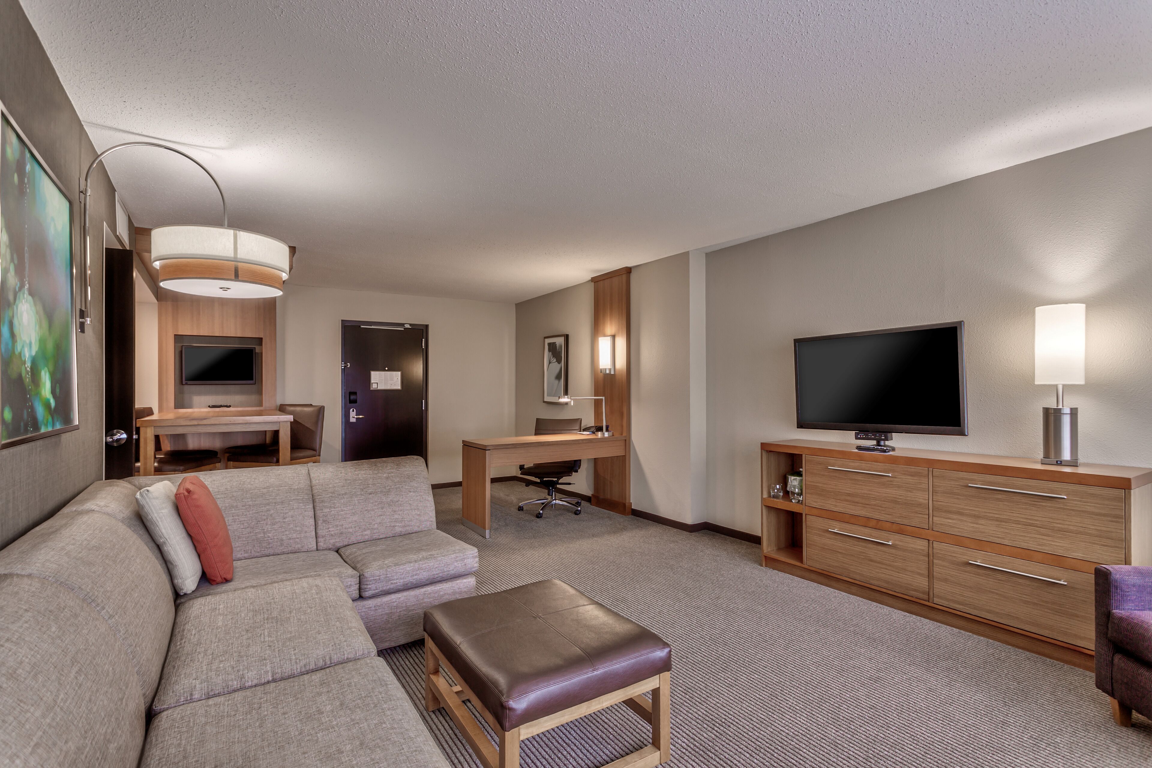 A hotel room with a sectional sofa, a small table, a desk with a chair, a television, and a cabinet.