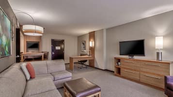 A hotel room with a sectional sofa, a small table, a desk with a chair, a television, and a cabinet.