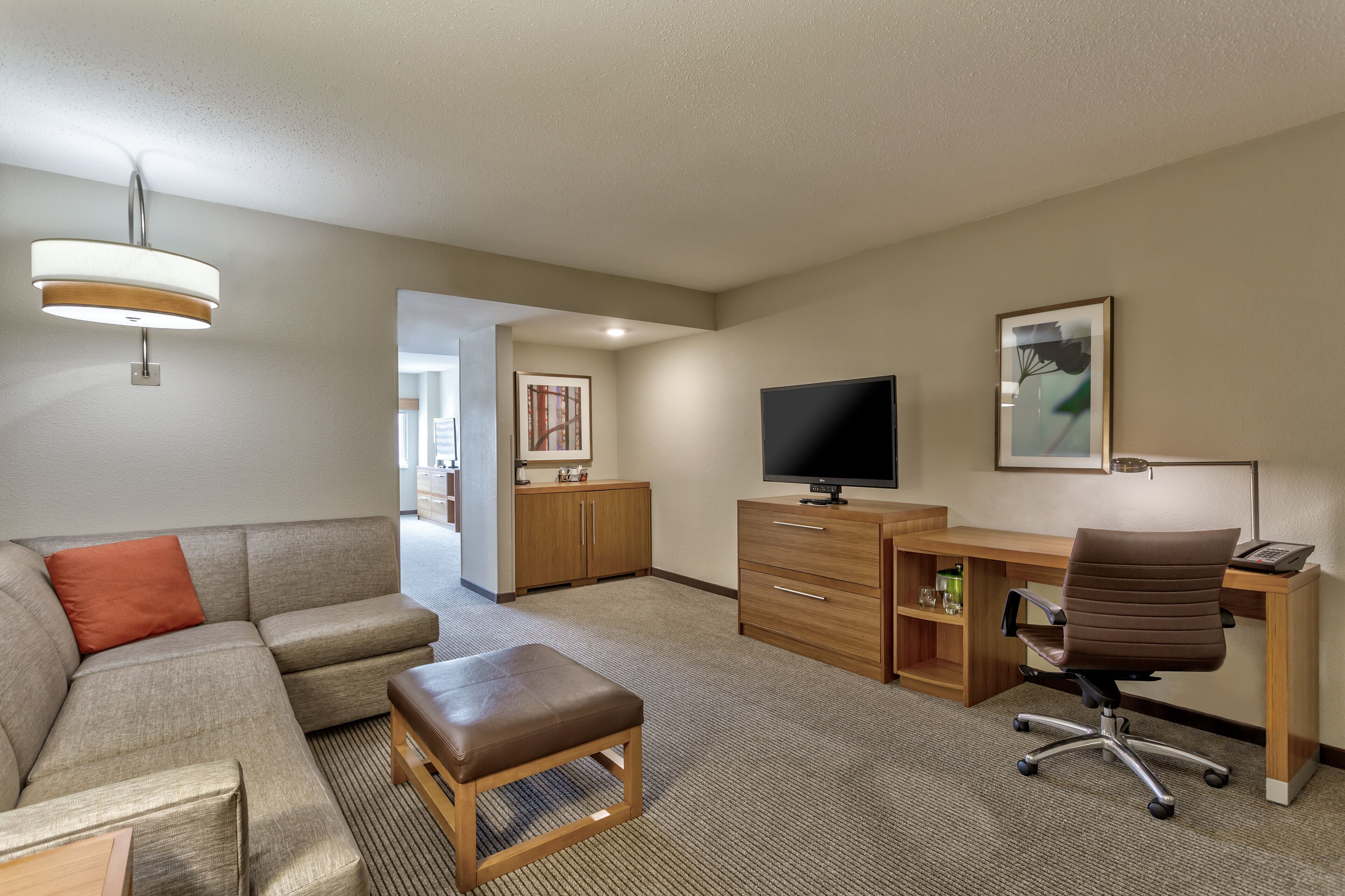 A hotel room with a sectional sofa, a wooden desk with a computer, a television, and a framed picture on the wall.