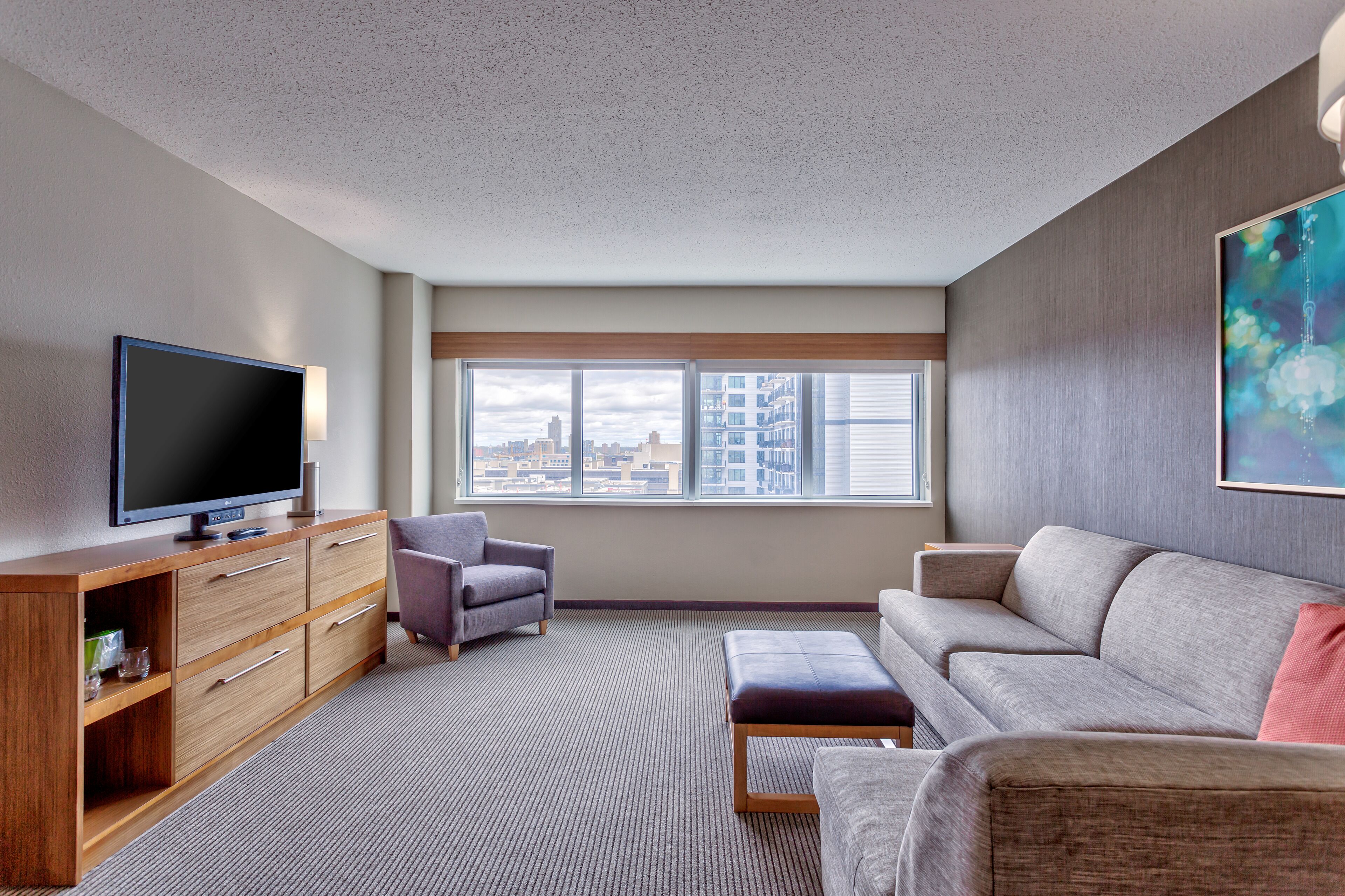 A modern living room with a flat-screen TV, a wooden cabinet, a grey sofa, and a large window with a city view.