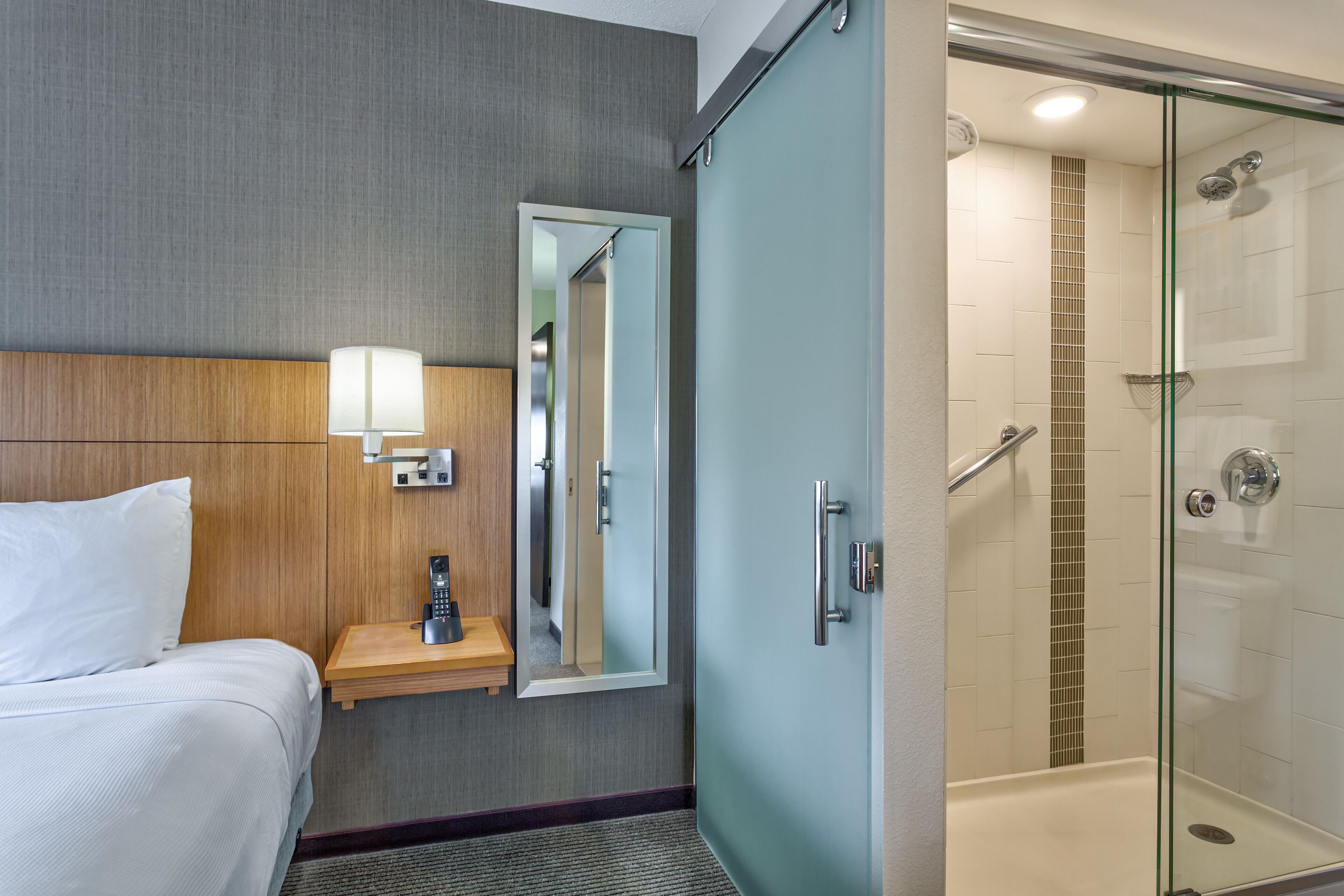 A hotel room with a bed, bedside lamp, mirror, and a glass-enclosed shower area.