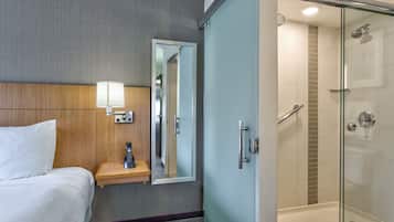A hotel room with a bed, bedside lamp, mirror, and a glass-enclosed shower area.
