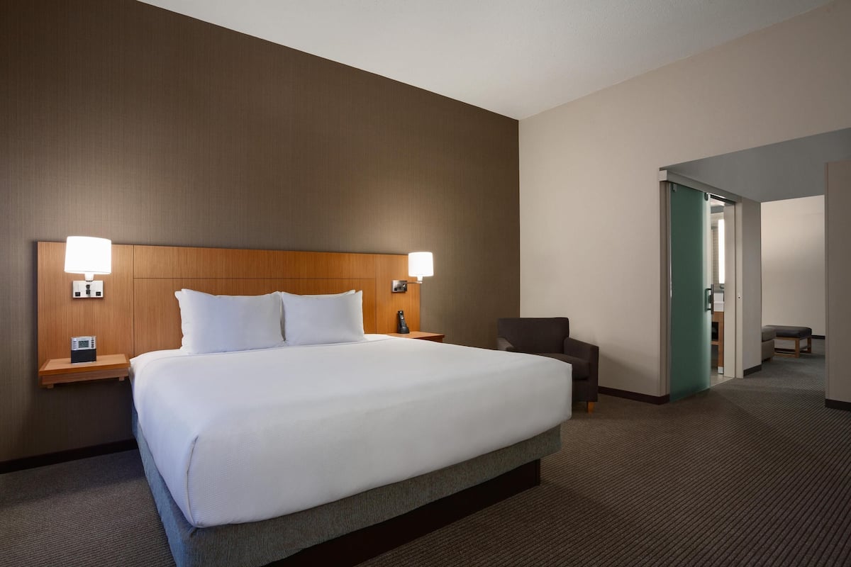 Een hotelkamer met een groot bed, twee nachtkastjes met lampen, een stoel en een deur die naar een andere kamer leidt.