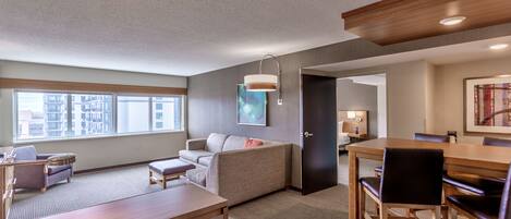 A modern hotel room with a large window, a sofa, a dining table, and a wooden ceiling.