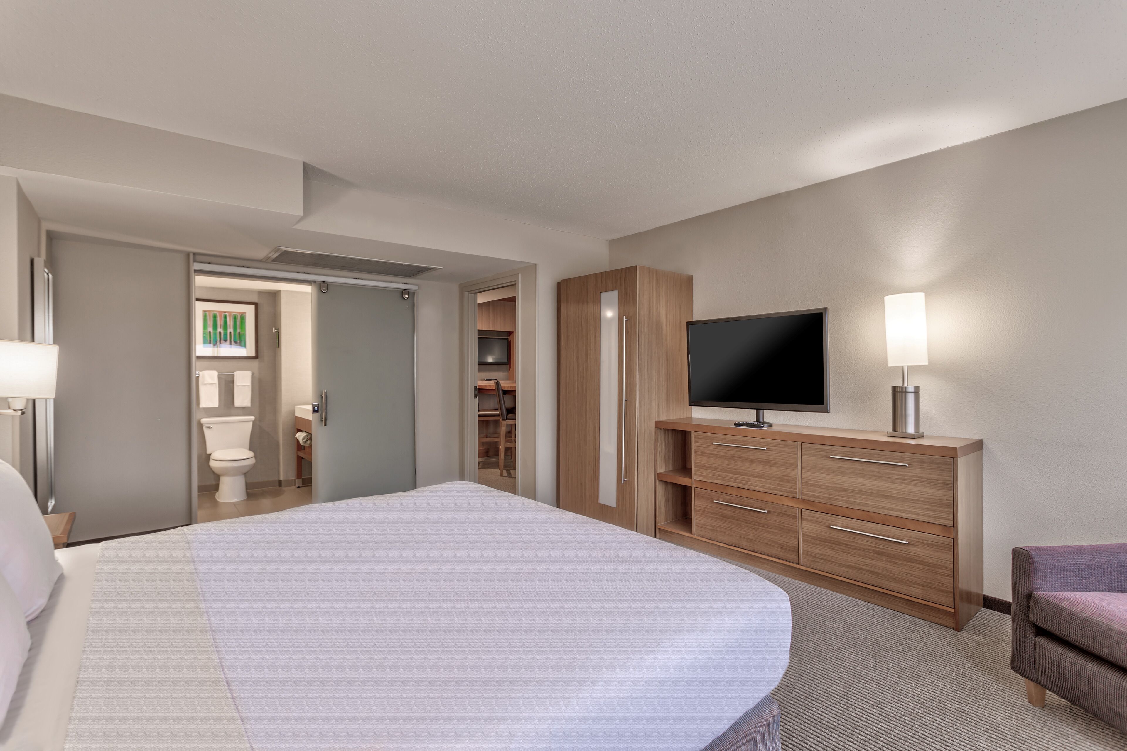 Une chambre d’hôtel avec un grand lit, un téléviseur posé sur une commode en bois, une lampe et une armoire avec des étagères.