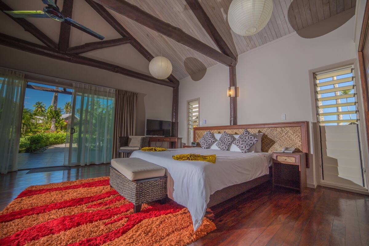 A bedroom with a large bed, a bedside table, a nightstand, a window with blinds, a ceiling fan, and a view of the outdoors.