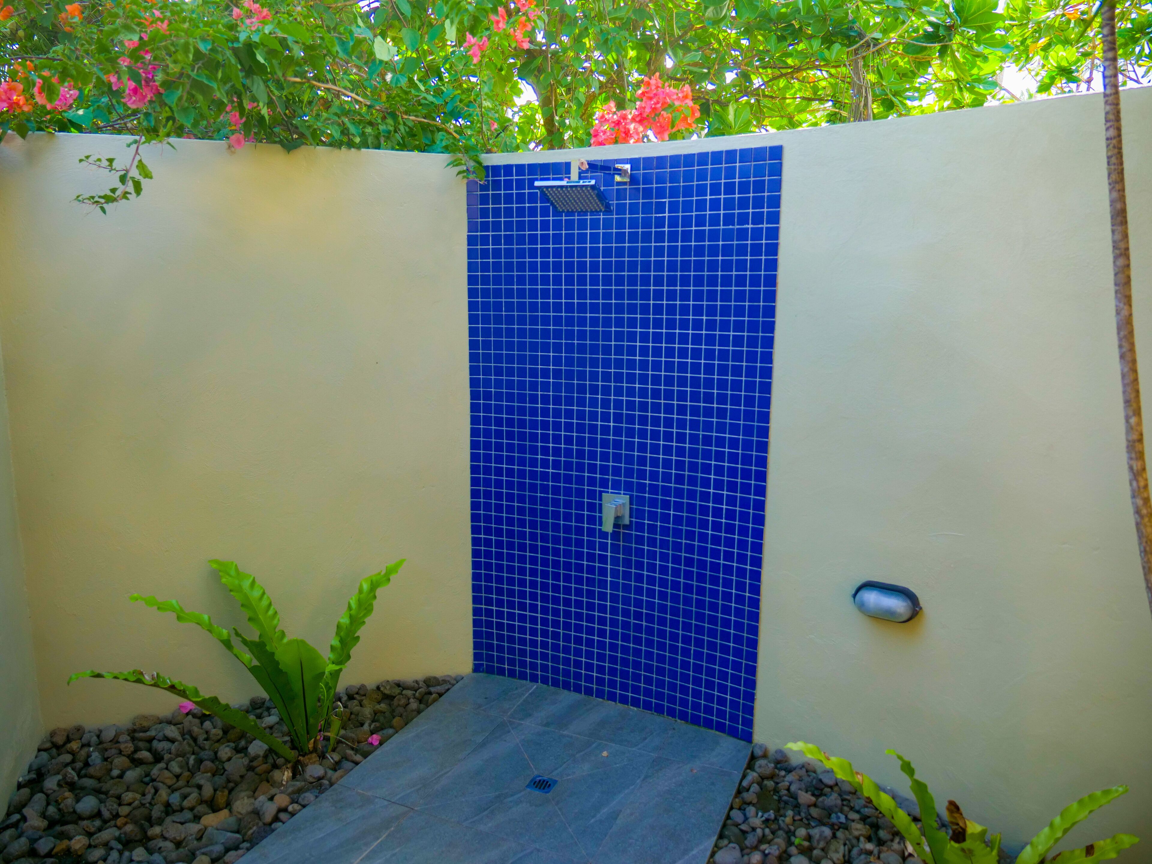 A small, enclosed outdoor shower area with a blue tiled wall, surrounded by plants and pebbles.