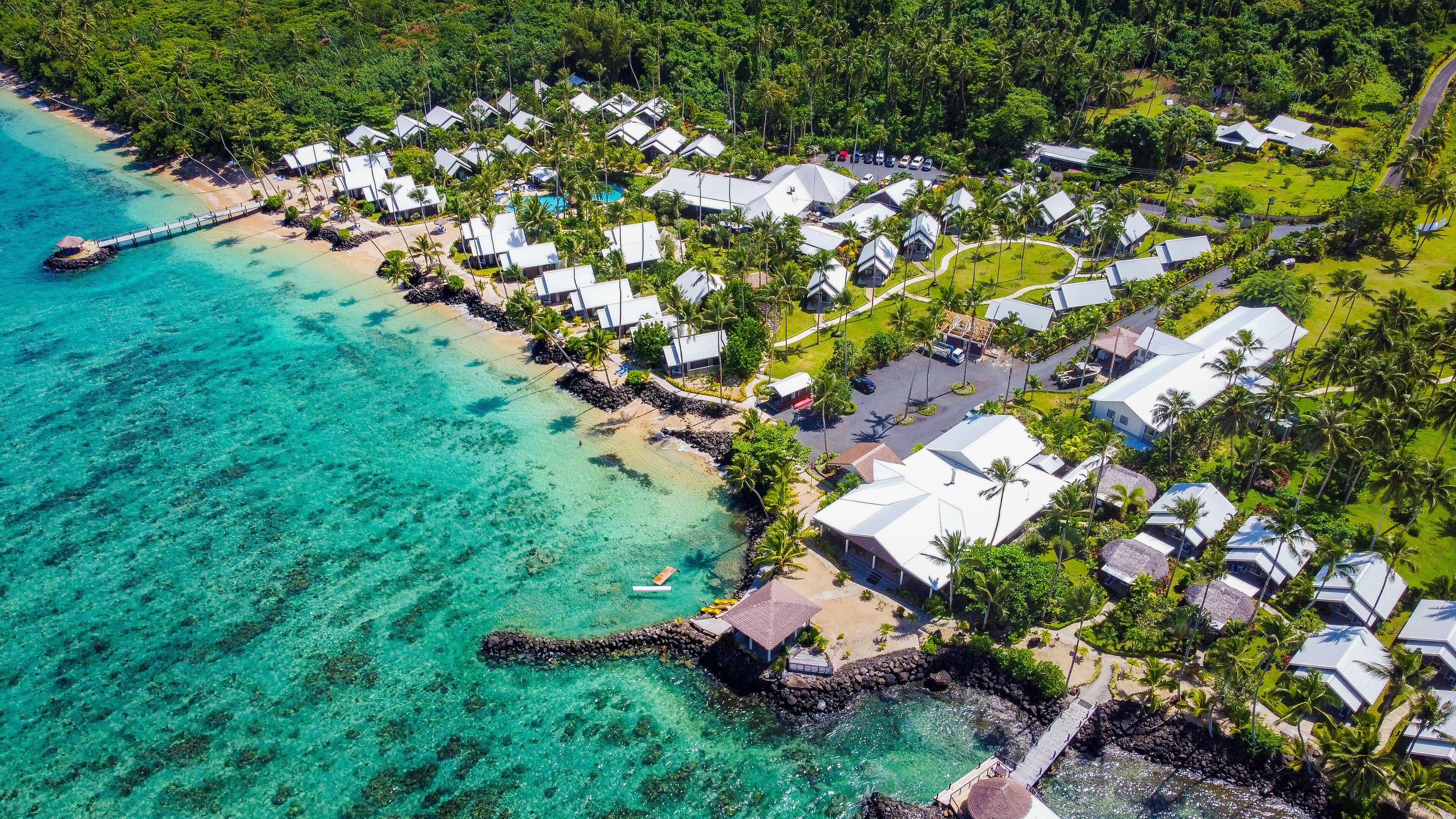 An aerial view of a coastal resort with bungalow-style accommodations, clear turquoise waters, and a rocky shoreline.
