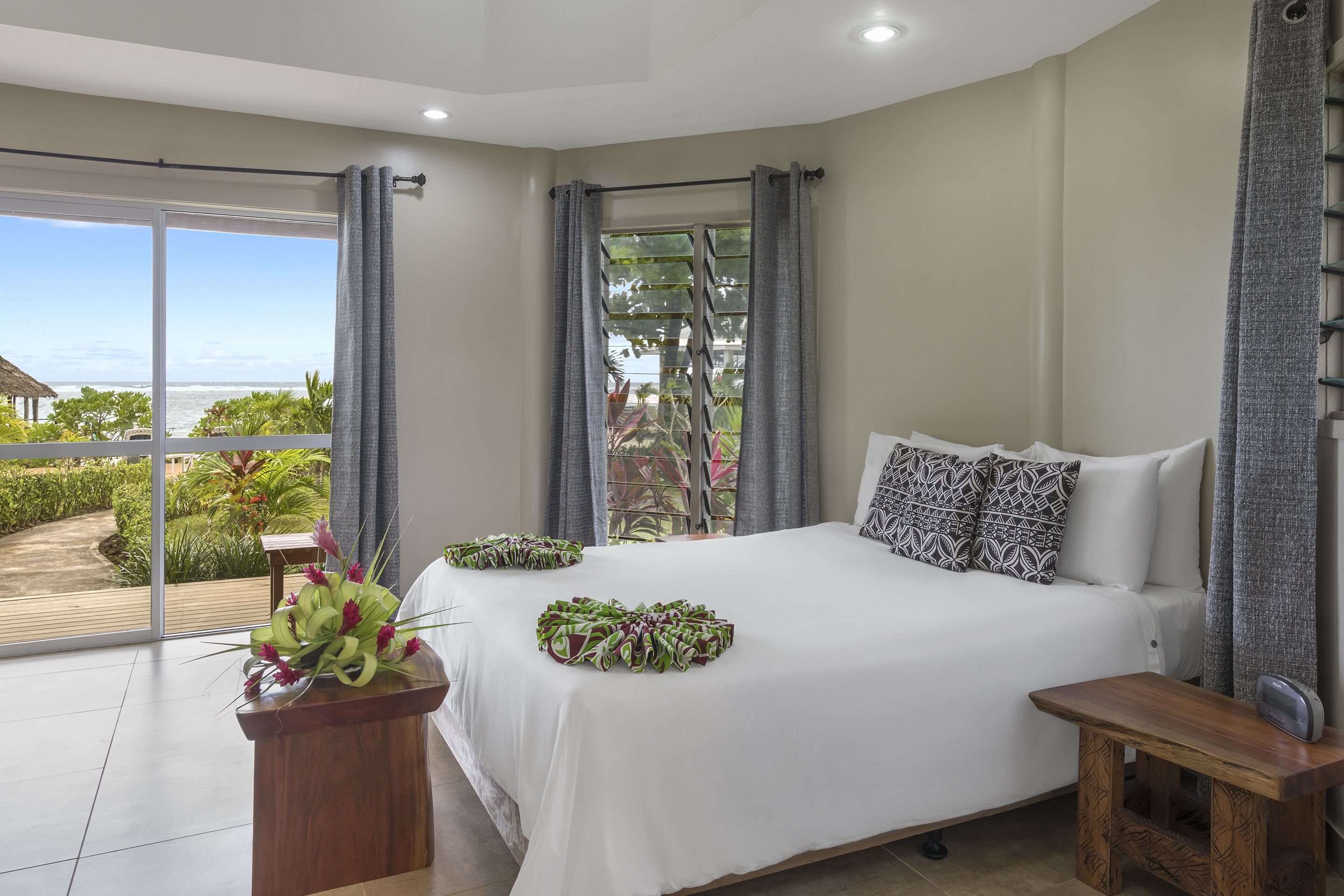 A bedroom with a large bed, a wooden nightstand, and a view of the ocean through a sliding glass door.