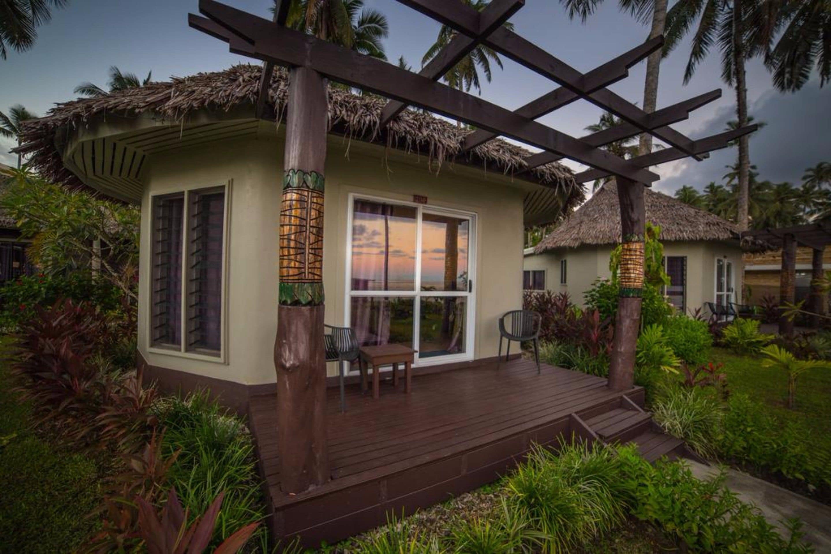Una cabaña con techo de paja, terraza de madera y jardín con plantas tropicales.