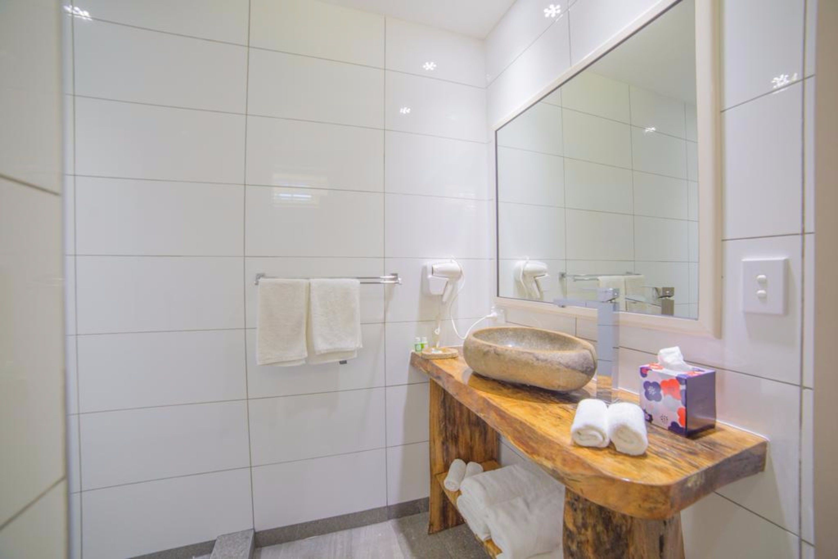 Un bagno moderno rivestito di piastrelle bianche, con un mobile lavabo in legno, un ampio specchio e cabina doccia in vetro.