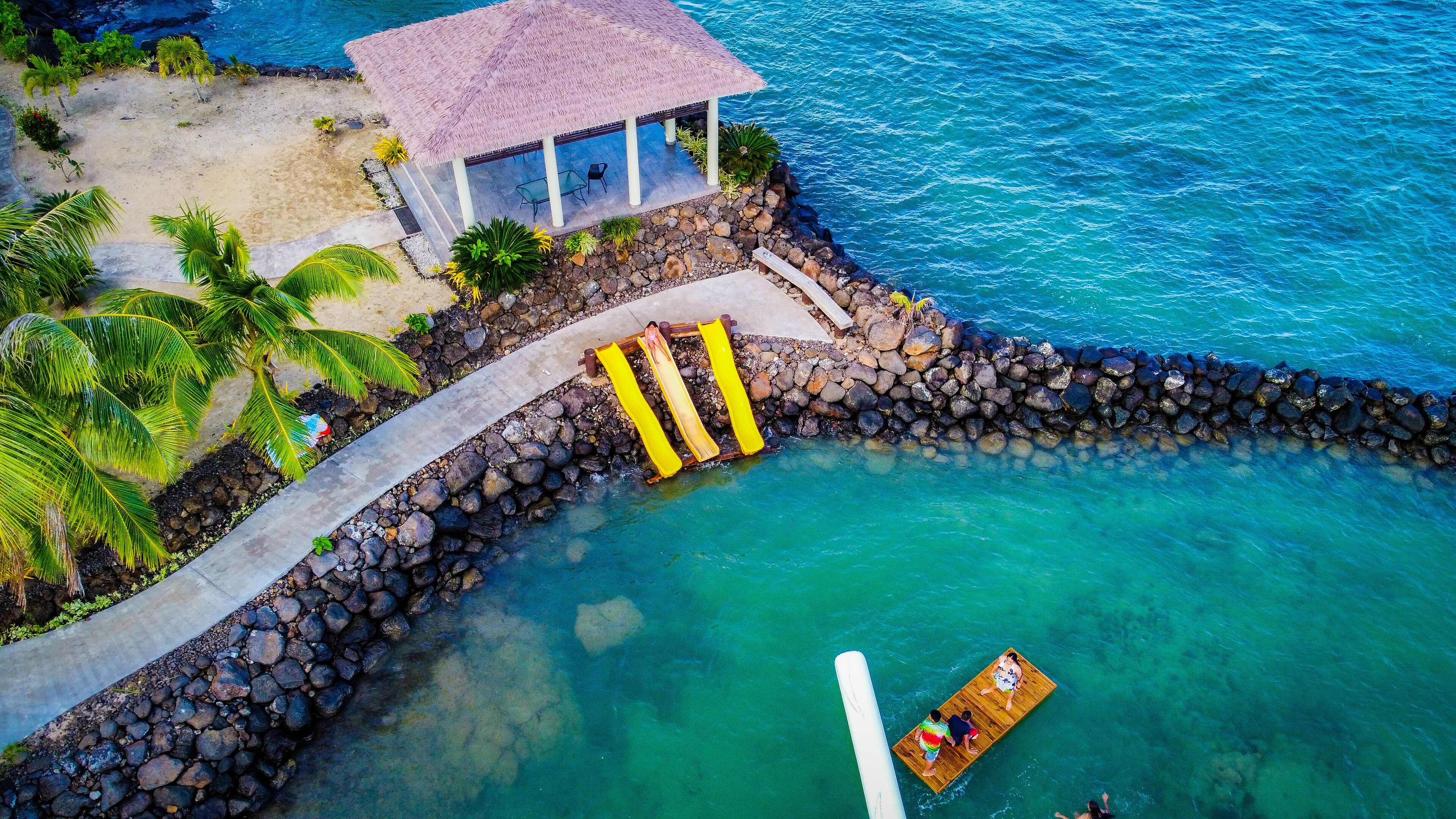 An aerial view of a beachside resort with a thatched-roof pavilion, a wooden boat dock, and a rocky shoreline.