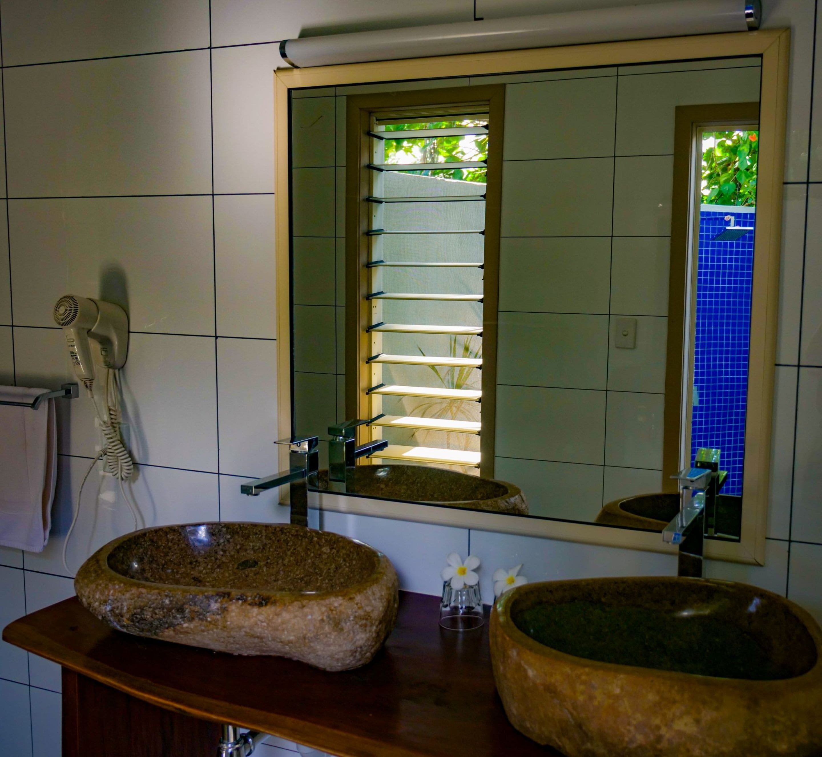 Baño con un espejo grande, dos lavamanos con lavabos de piedra natural, un perchero y una ventana con persianas.