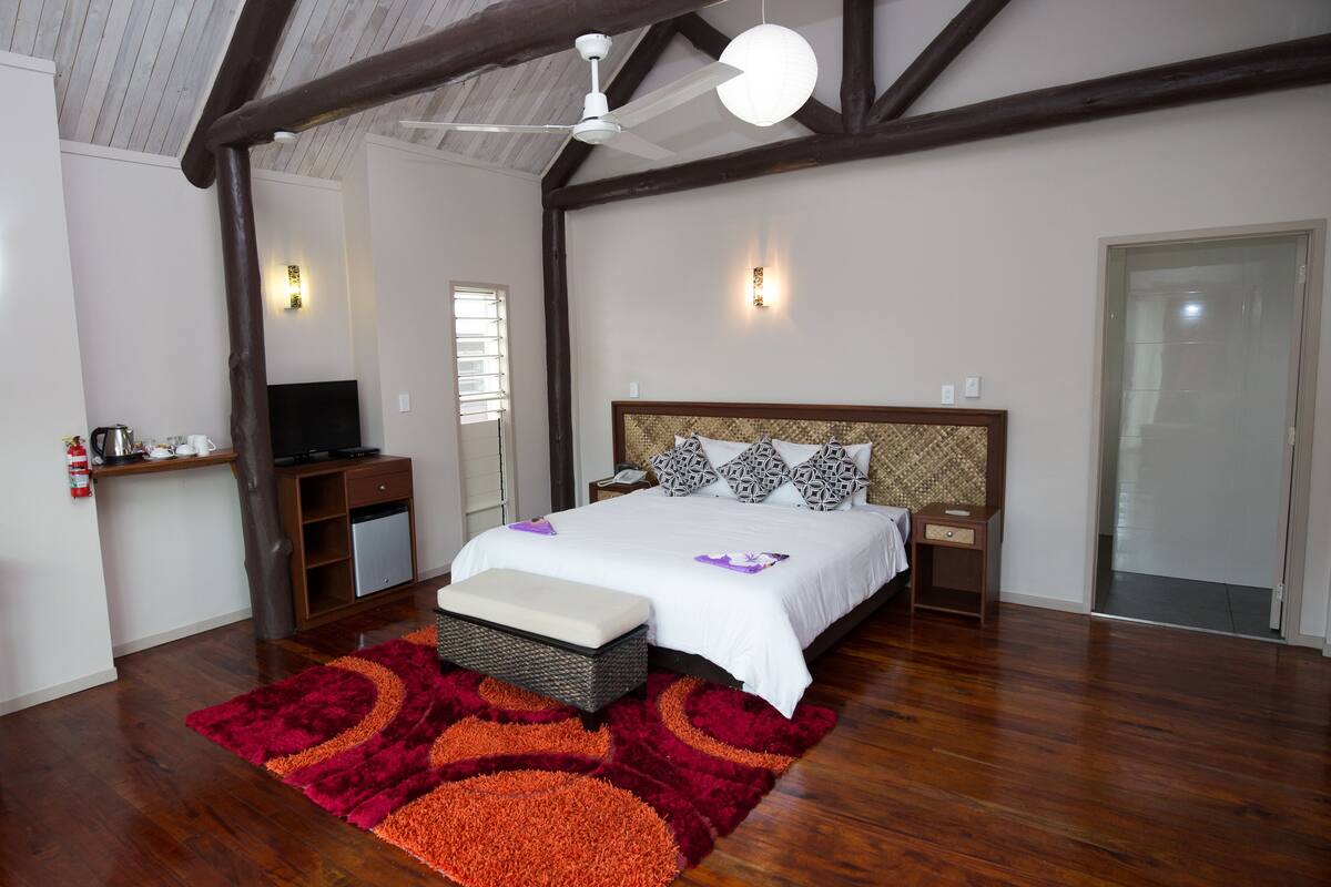 A bedroom with a large bed, wooden beams, a ceiling fan, a TV, and a red and orange rug.