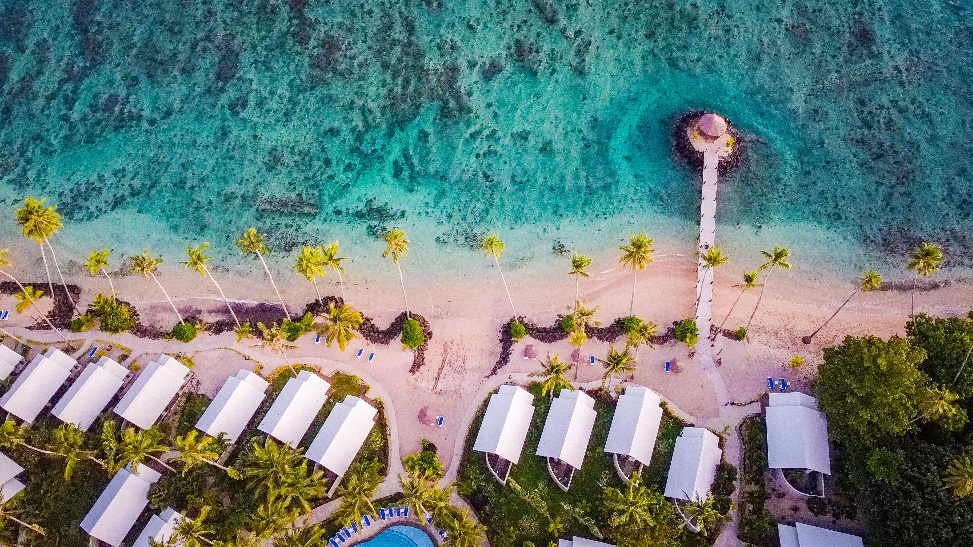 Vista aérea de un complejo turístico en la playa con filas de cabañas blancas, palmeras y un mar turquesa transparente.