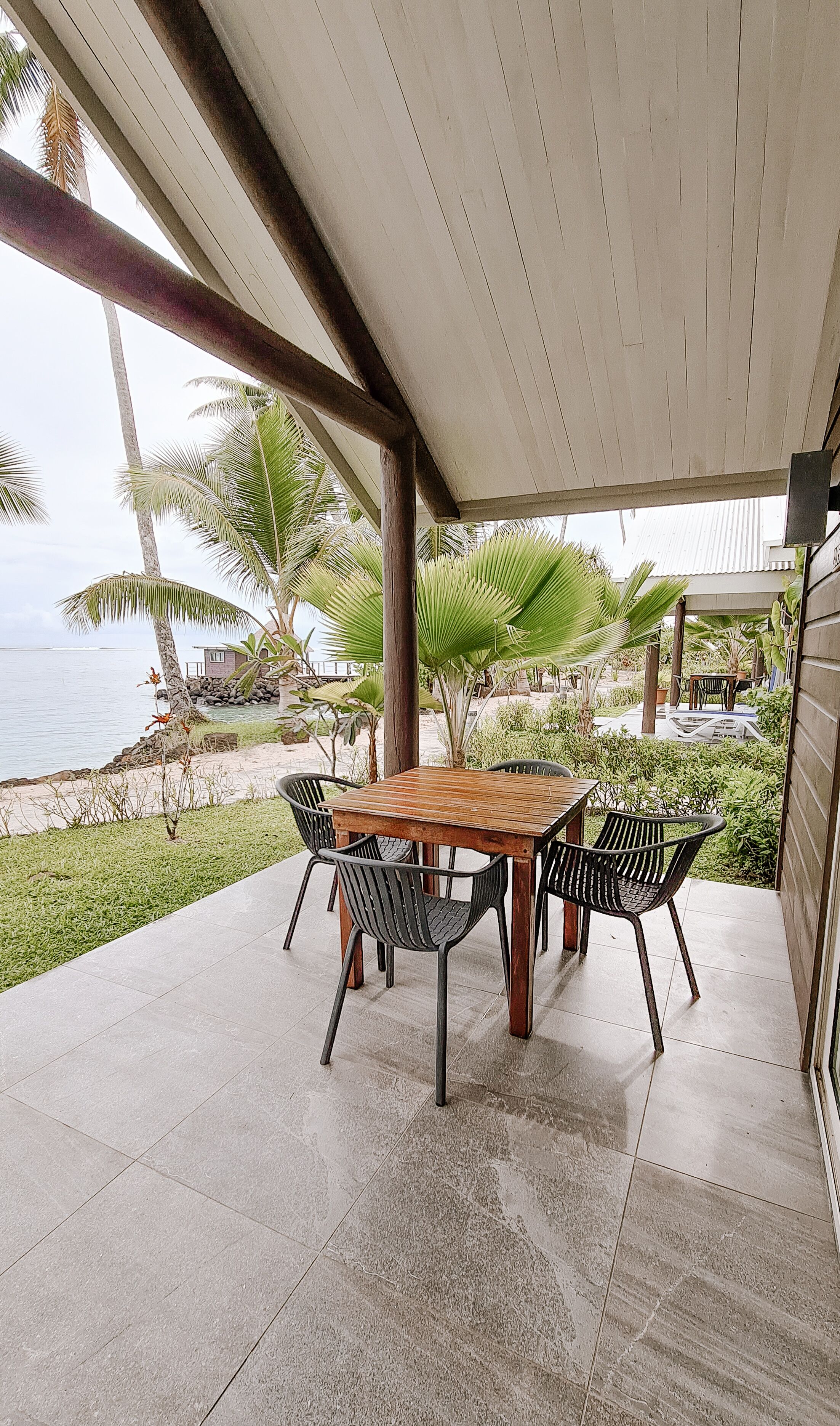 An outdoor patio with a wooden table and metal chairs, overlooking a beach with palm trees and a view of the ocean.