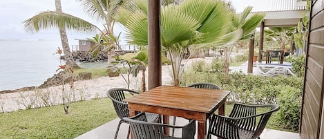 An outdoor patio with a wooden table and metal chairs, overlooking a beach with palm trees and a view of the ocean.