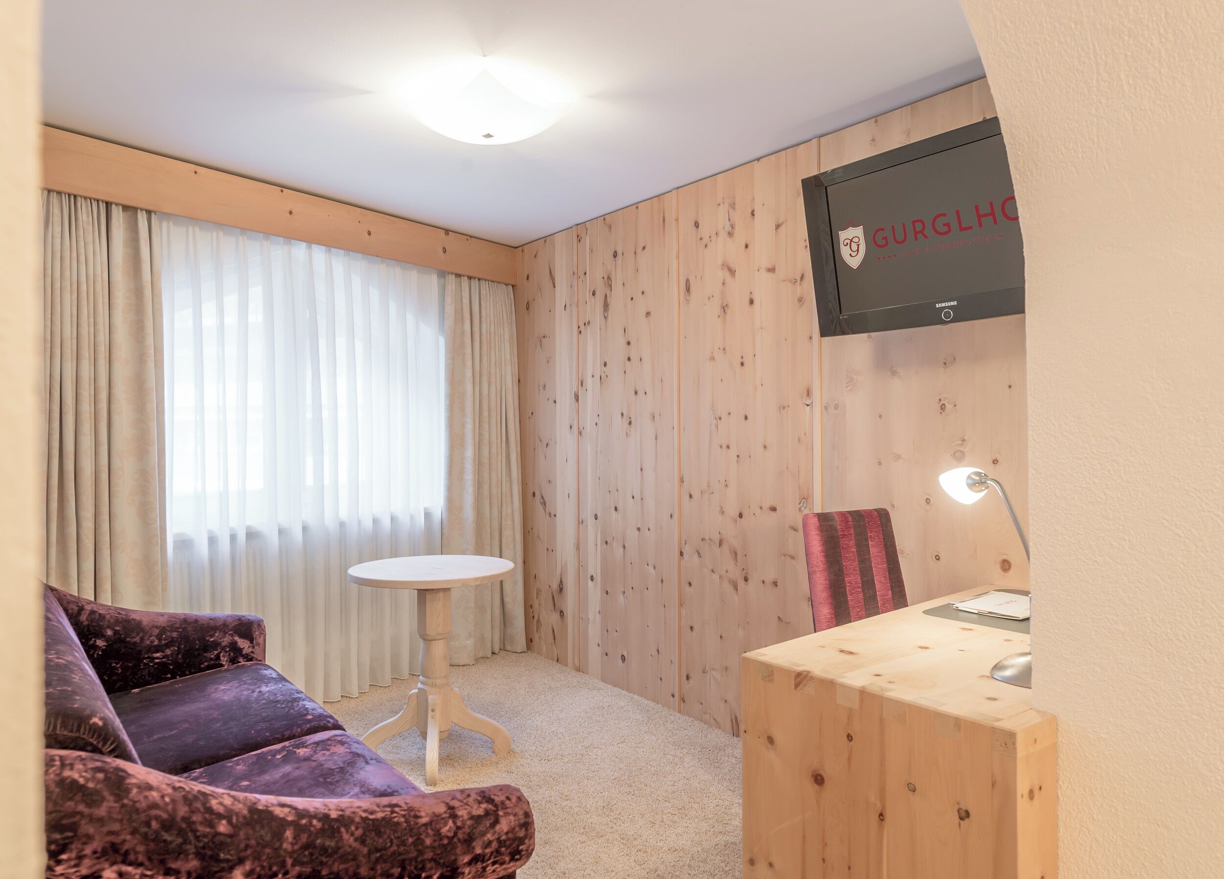 A hotel room with a wooden panel wall, a TV mounted on the wall, a small round table, a purple sofa, and a desk with a lamp.