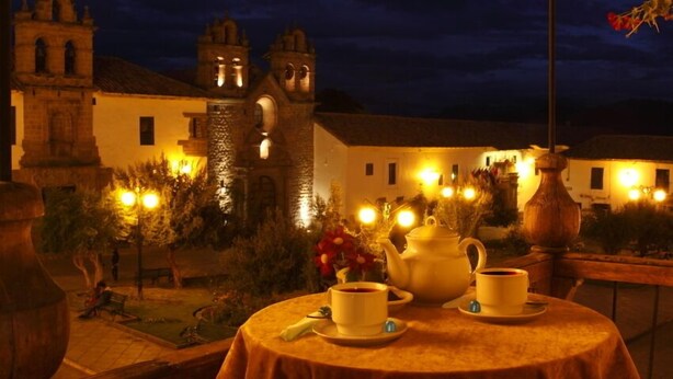 An outdoor dining area with a table set for two, surrounded by a garden and historic buildings in the background. - Fallen Angel (Cusco)