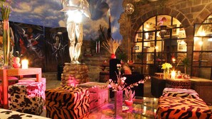 A cozy lounge area with plush, patterned seating, a decorative winged figure, and a stone wall backdrop. - Fallen Angel (Cusco)