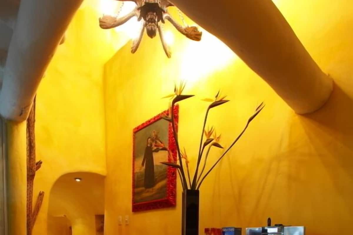A room with a wooden floor, a desk with a patterned tablecloth, a chair, a vase with branches, a framed painting, and a ceiling fan.