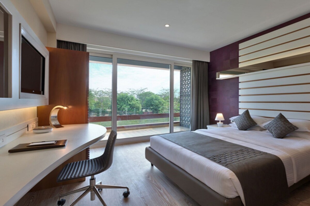 A modern hotel room with a large bed, a desk, and a view of greenery outside.