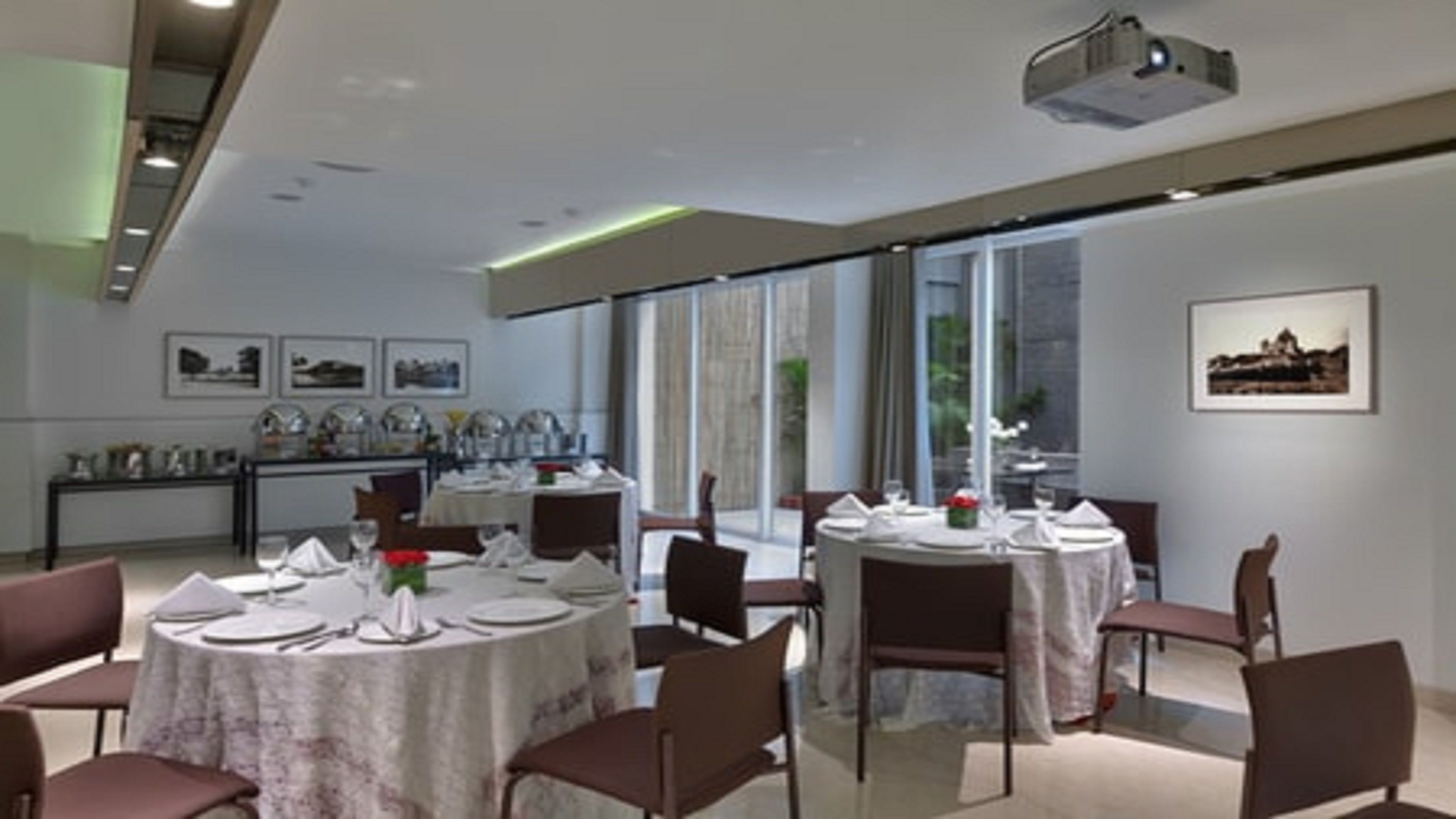 Een moderne eetruimte met ronde tafels voor vier personen, elk met een witte tafelkleed en rode bloemen. Er staan bruine stoelen bij de tafels. De kamer heeft grote ramen met gordijnen, waardoor natuurlijk licht binnenkomt.