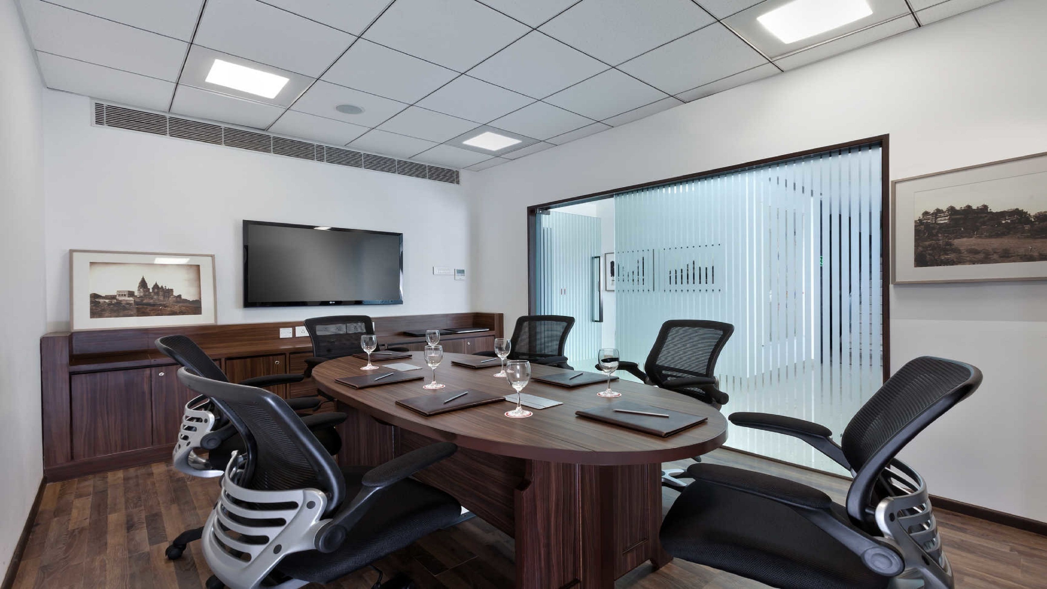 A conference room with a large table, four chairs, a flat-screen TV, and a large window with blinds.