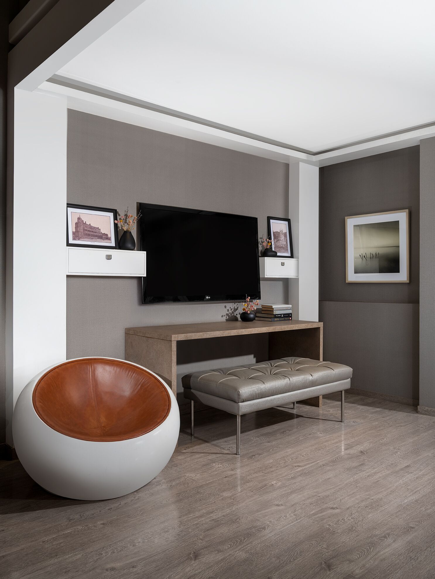 A modern living room with a large flat-screen TV, a round leather chair, a cushioned bench, and framed pictures on the walls.