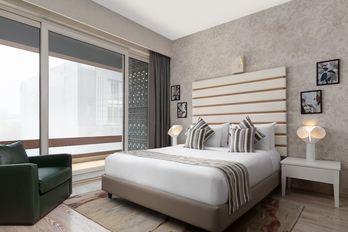 A modern hotel room with a large bed, a green armchair, a nightstand with a lamp, and a view of the city through large windows.