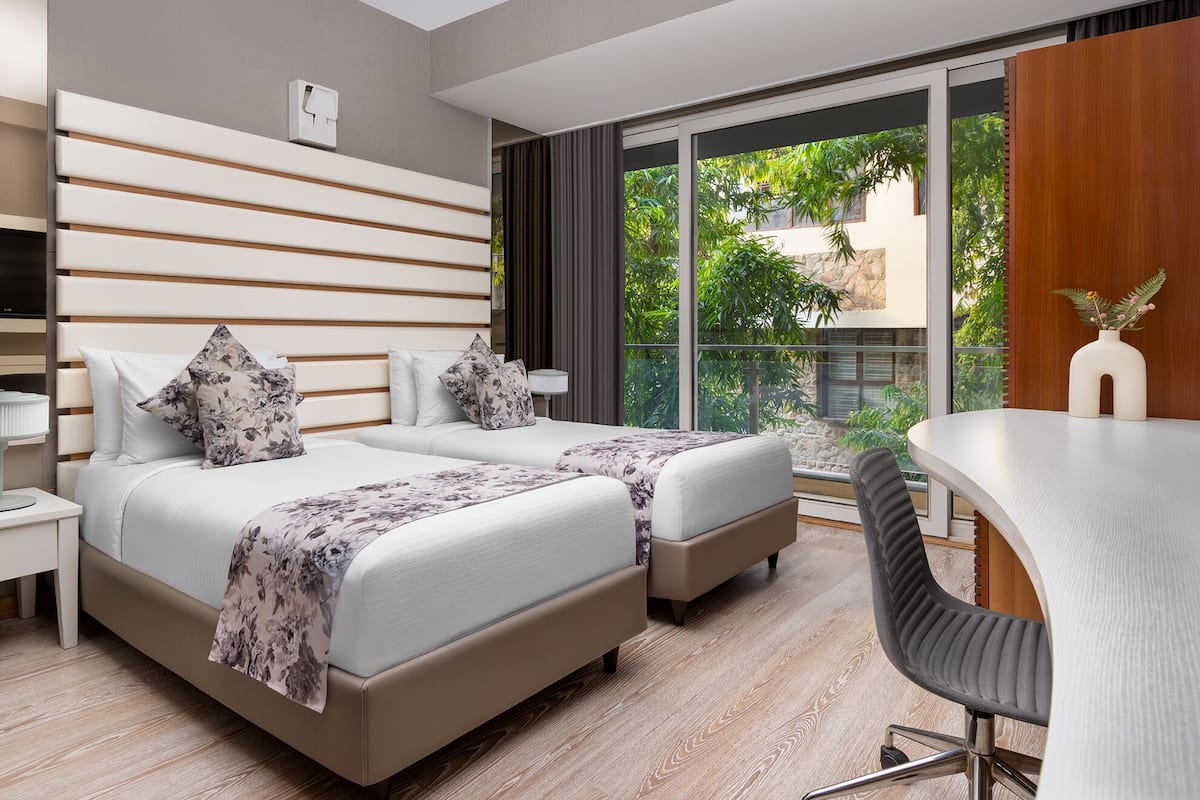 A modern hotel room with two beds, a desk, and a large window offering a view of greenery.
