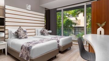 Una habitación de hotel moderna con dos camas, un escritorio y un amplio ventanal que ofrece vistas a zonas verdes.