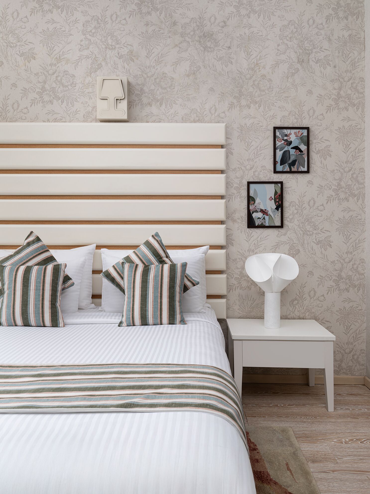 Un dormitorio con colcha a rayas, almohadas blancas, cabecero de madera, mesita de noche blanca con una lámpara y papel tapiz con motivos florales.